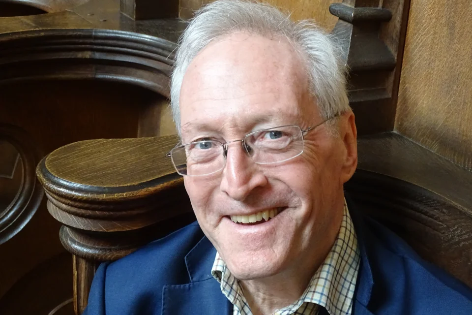 Headshot of Reverend Professor Diarmaid MacCulloch, wearing a navy suit jacket and glasses, sitting in a church pew.