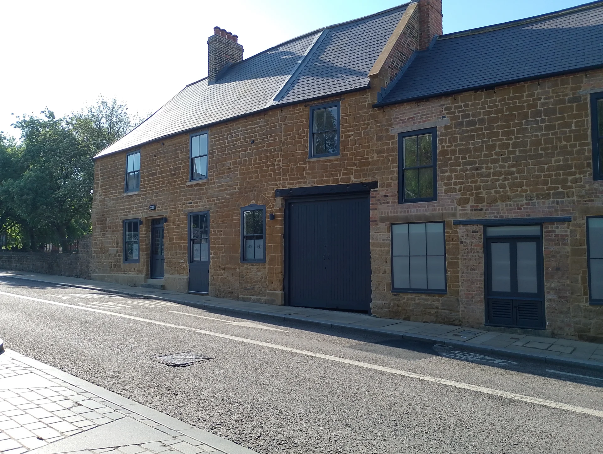 A photograph of a historic pub sat on a main road. The street is empty. It's a sunny day. The pub lies in the share, but the street lies in the sun. The pub is built from orange and yellow Northamptonshire ironstone and has a black slate roof. It is closed, there is no activity going on.