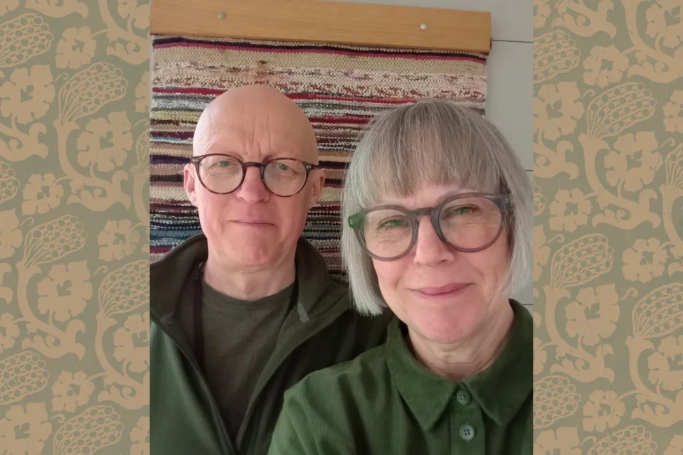 A woman with glasses and neat grey bob and green blouse in the foreground smiling. A bald man with glasses behind her.