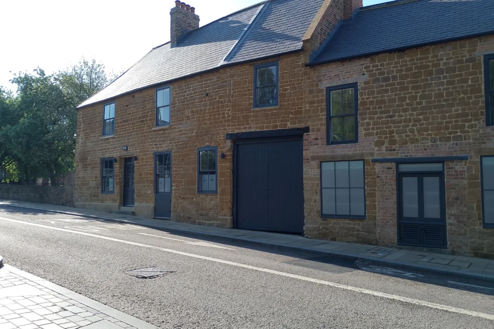 A photograph of a historic pub sat on a main road. The street is empty. It's a sunny day. The pub lies in the share, but the street lies in the sun. The pub is built from orange and yellow Northamptonshire ironstone and has a black slate roof. It is closed, there is no activity going on.
