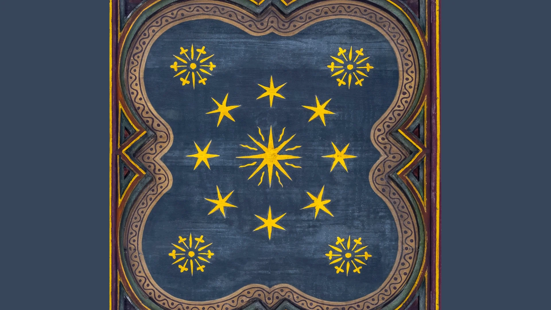 a photograph of a ceiling motif. A large yellow star is surrounded by 8 smaller stars. In each corner there are yellow snowflakes, all bordered by a decretive design.