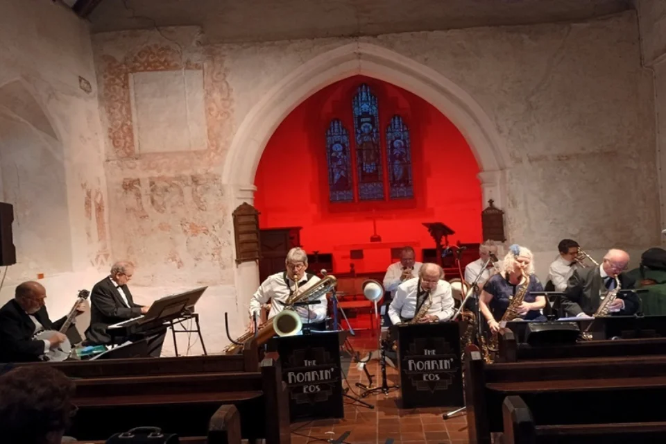 Jazz band plays in a church, musicians seated with instruments before a red-lit altar and stained glass window.