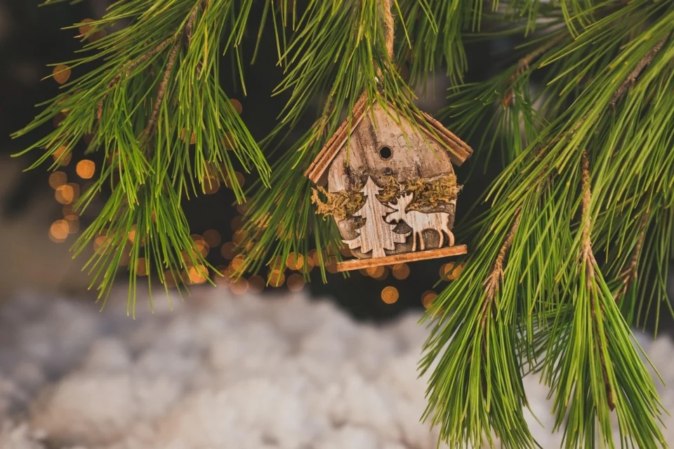 Small wooden birdhouse ornament hanging from pine branches, with festive lights softly glowing in the background.