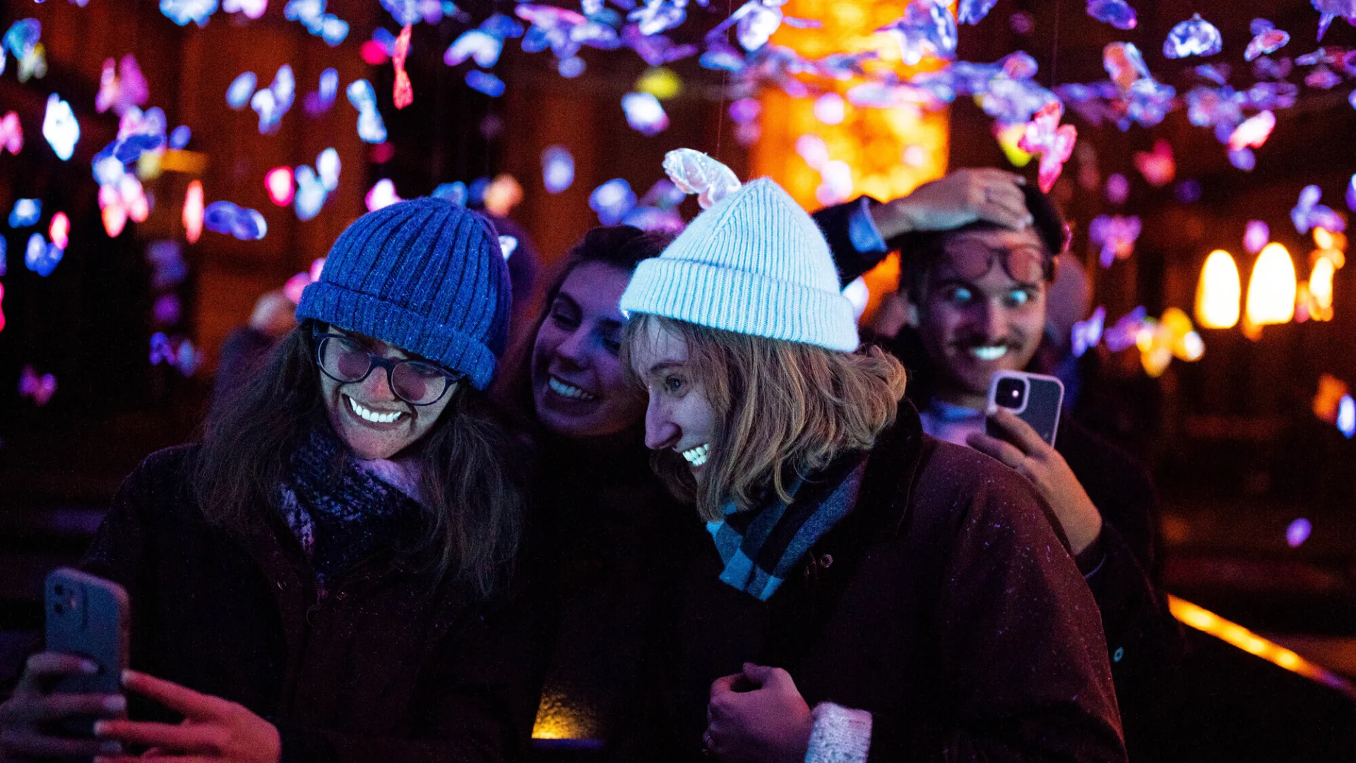 Two people lit up by blacklight and taking selfies in a church decorated with light-up butterflies.