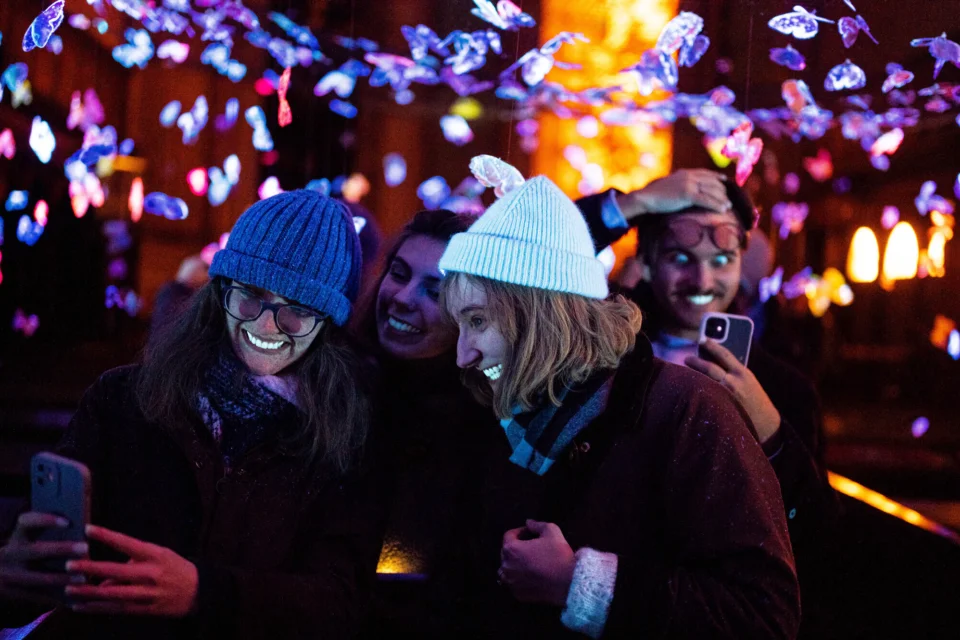 Two people lit up by blacklight and taking selfies in a church decorated with light-up butterflies.