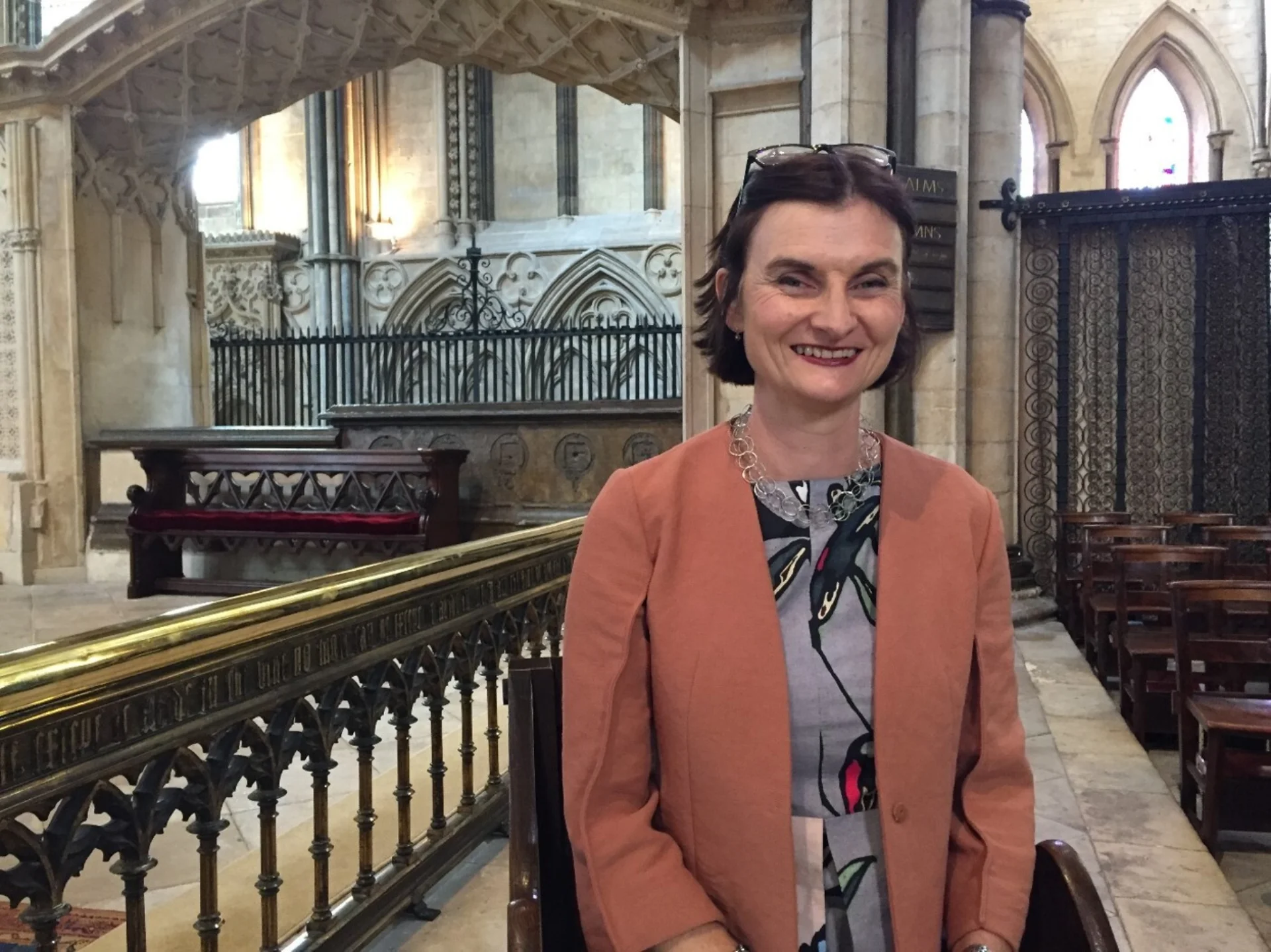 White woman with dark hair wearing grey dress and peach blazer stands inside stone church
