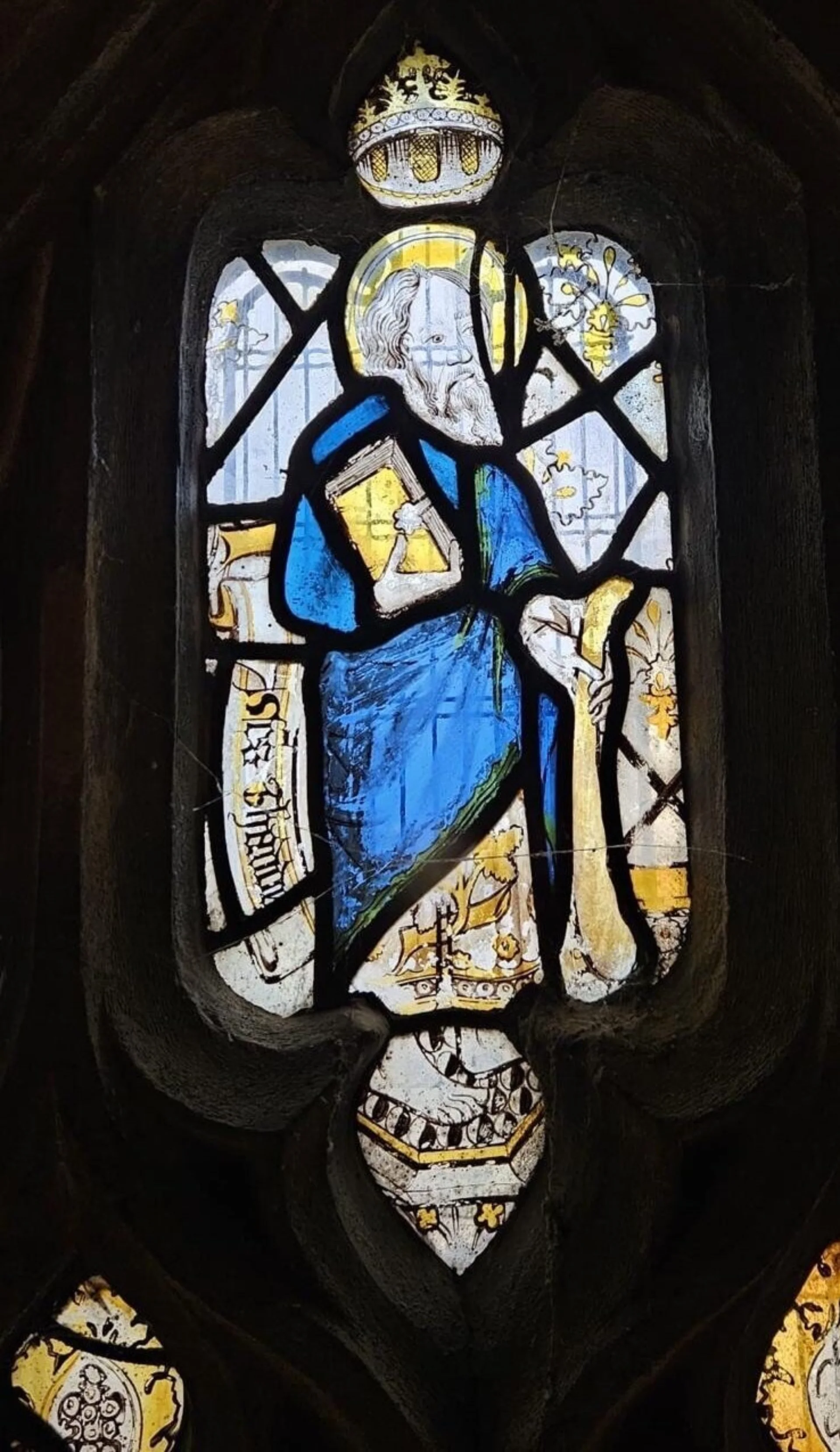 Medieval stained glass depicting a saint with a crown and a long beard, dressed in a blue cloak and holding a simple staff.