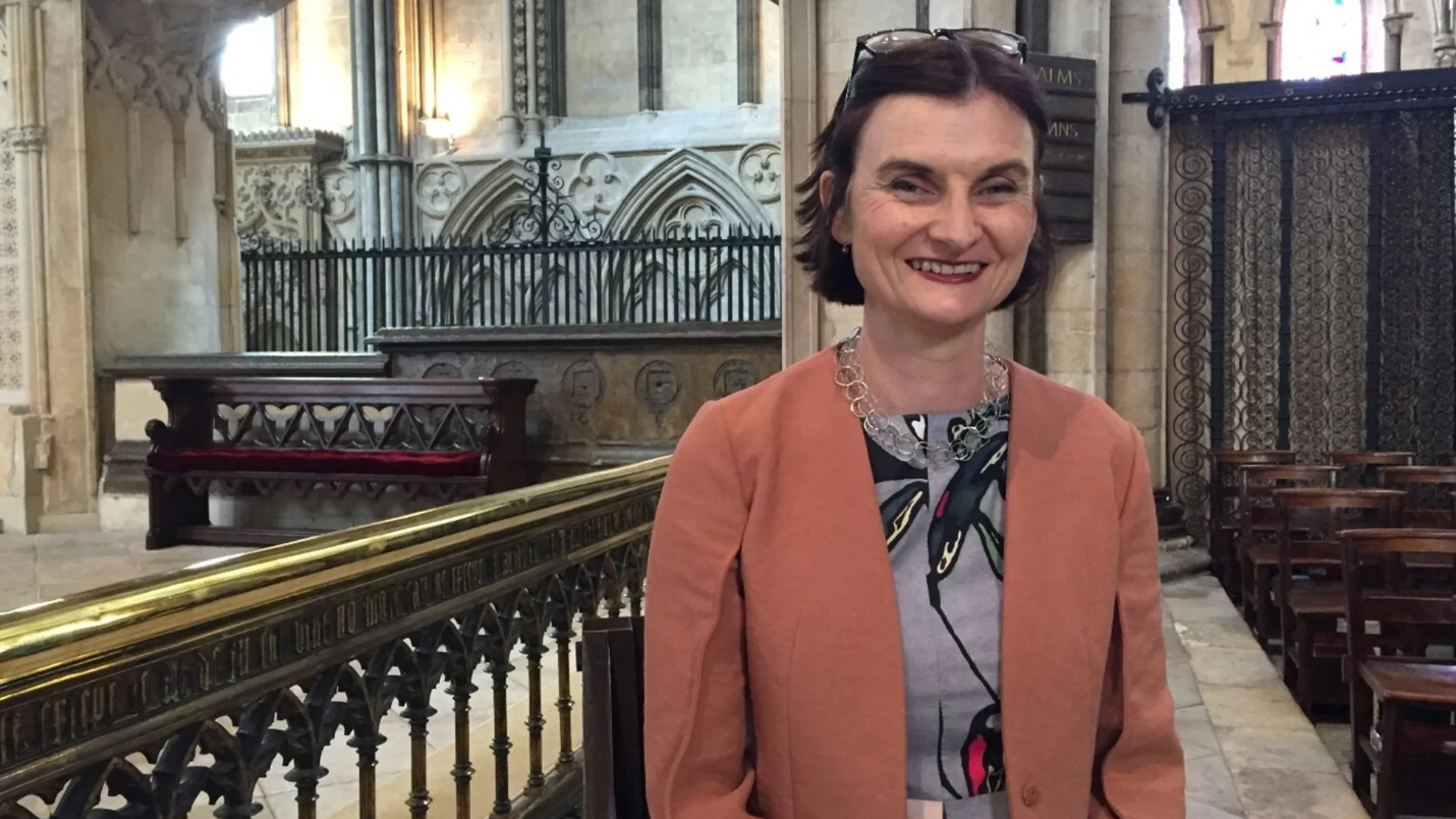 White woman with dark hair wearing grey dress and peach blazer stands inside stone church