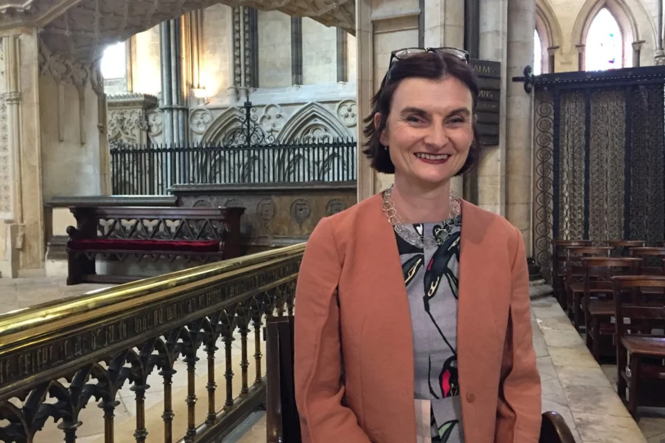 White woman with dark hair wearing grey dress and peach blazer stands inside stone church