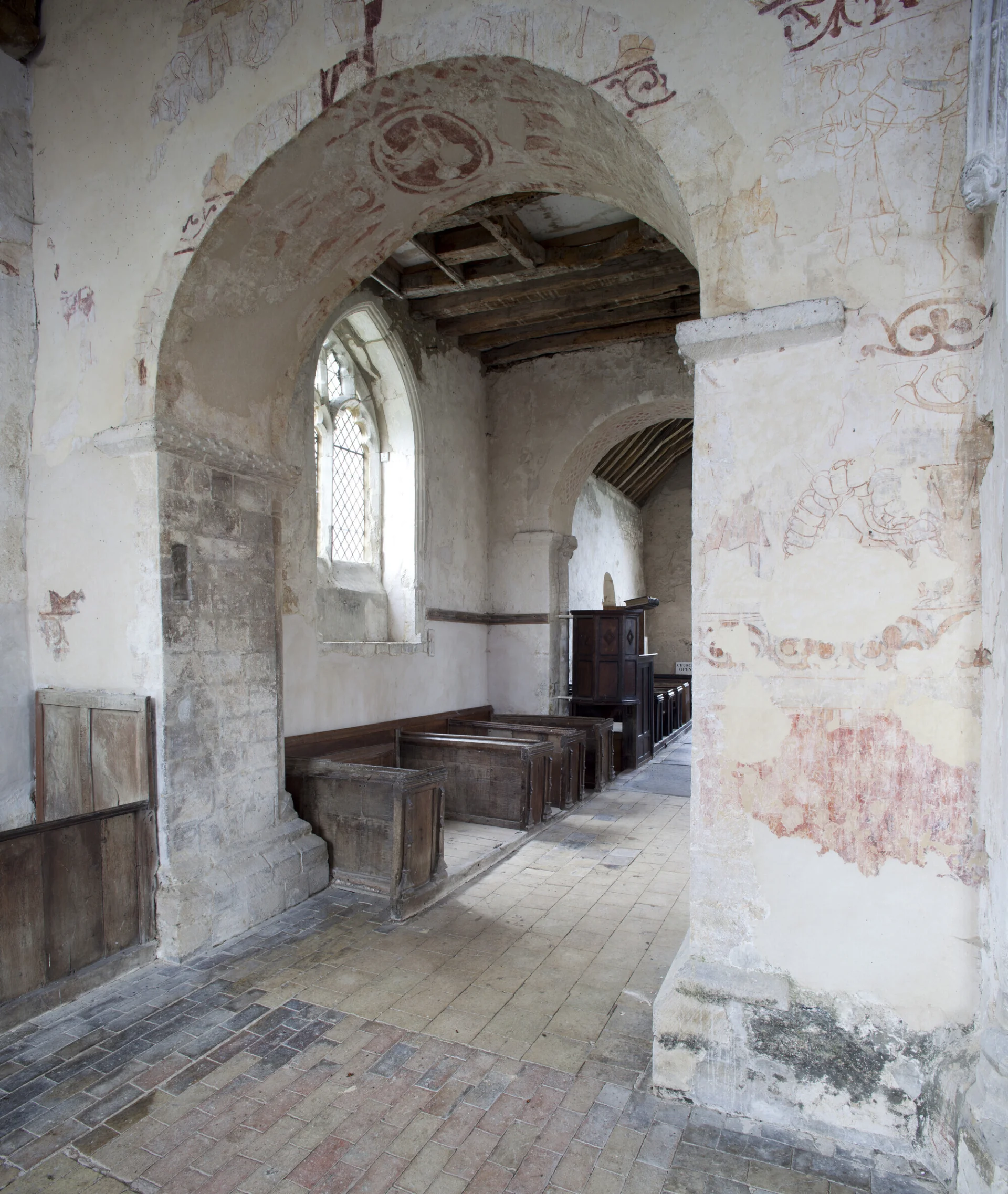 Decorative wall paintings in shades of red and yellow on a wall and arch at St. John's, Duxford, a medieval church in Cambridgeshire.