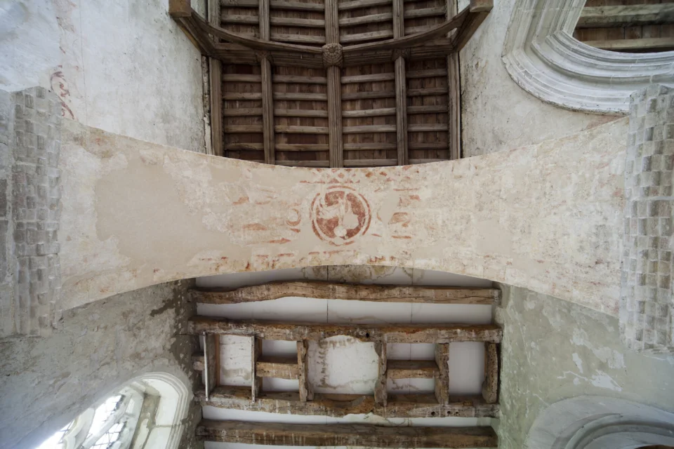 Photograph of a romanesque arch taken from below to show the remnants of decorative red, medieval paint patterns inside the arch. Above the arch we can see the old, warped beams of a wooden ceiling.